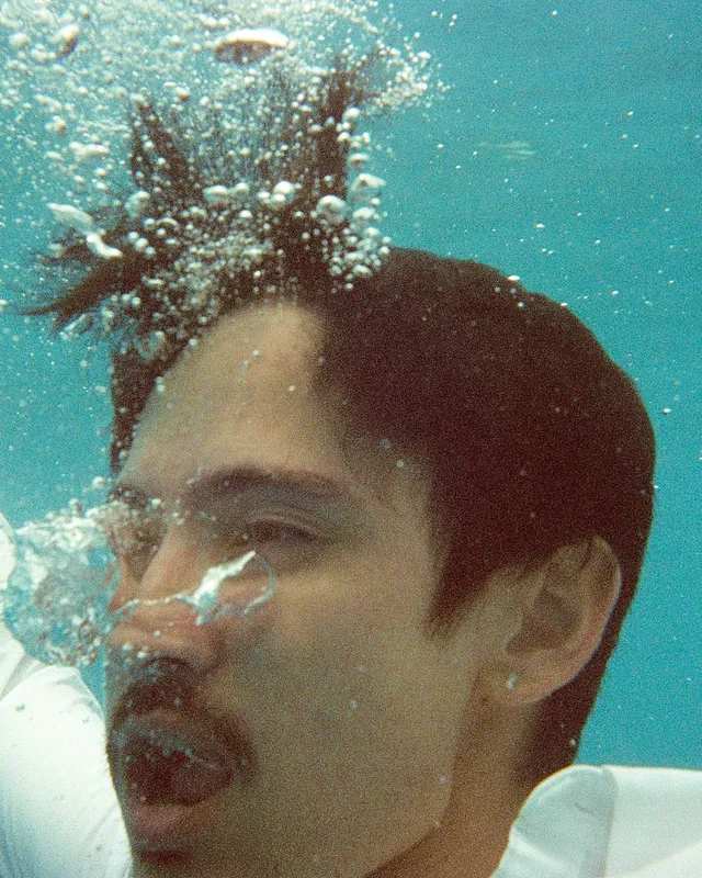 Mettā Atom submerged in turquoise water, exhaling a stream of bubbles