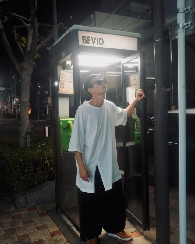 Mettā Atom looking skyward beside a phone booth at night, sunglasses resting on his cap